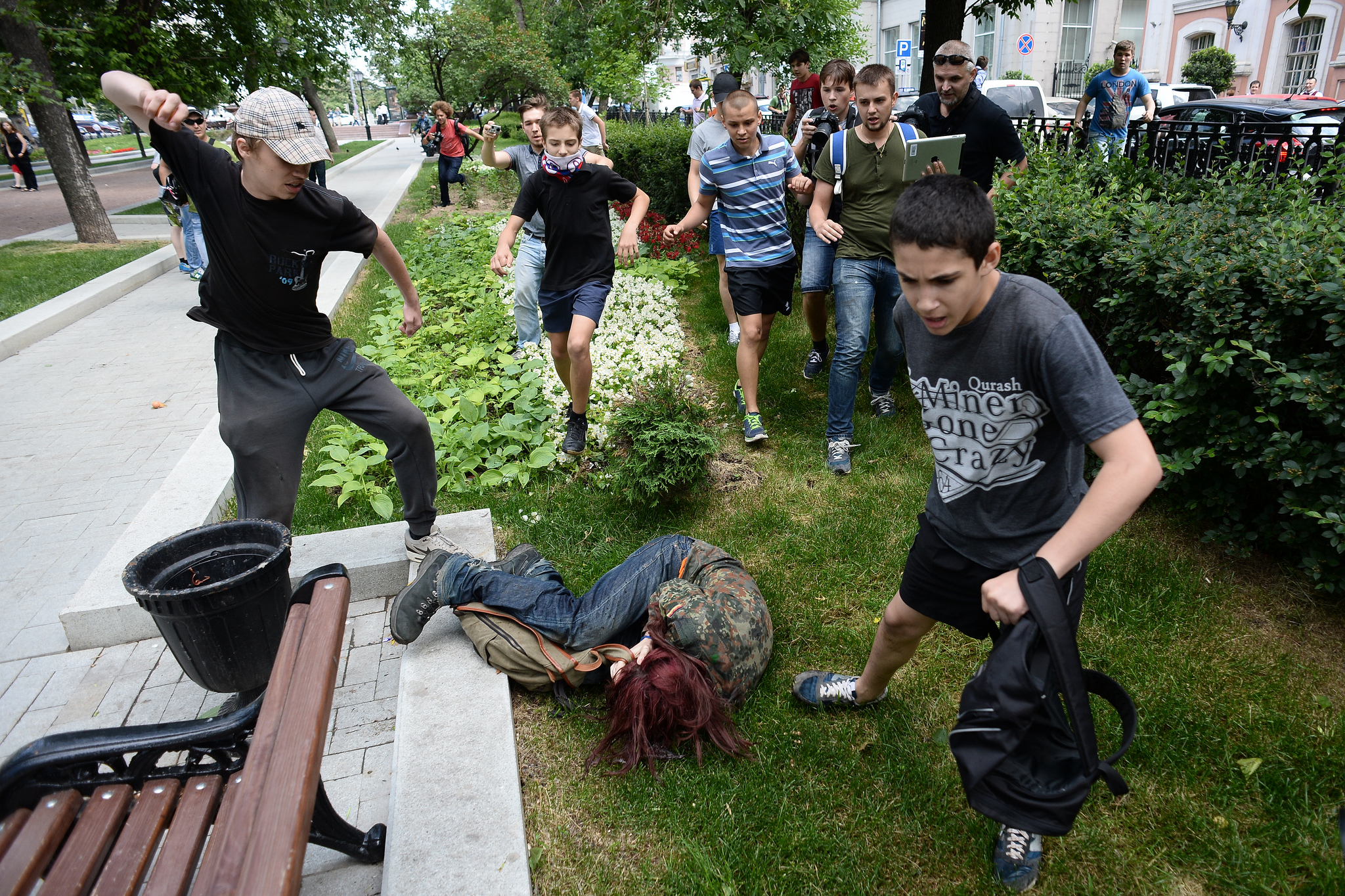 A teenager hits another teen who is curled up on the ground, a large group of teens approach.