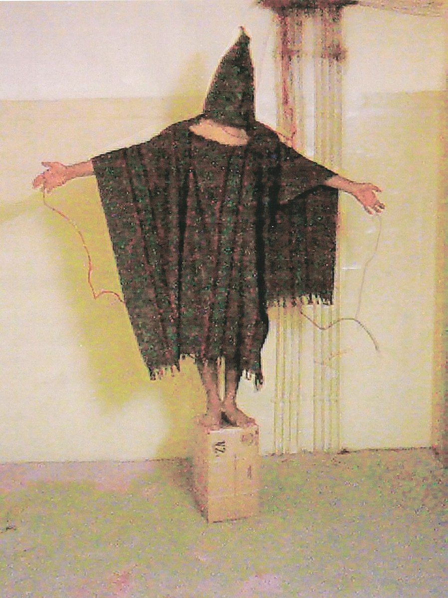 A man standing on a box with wires connected to his hands, wearing only a long poncho with a pointed hood covering his face.