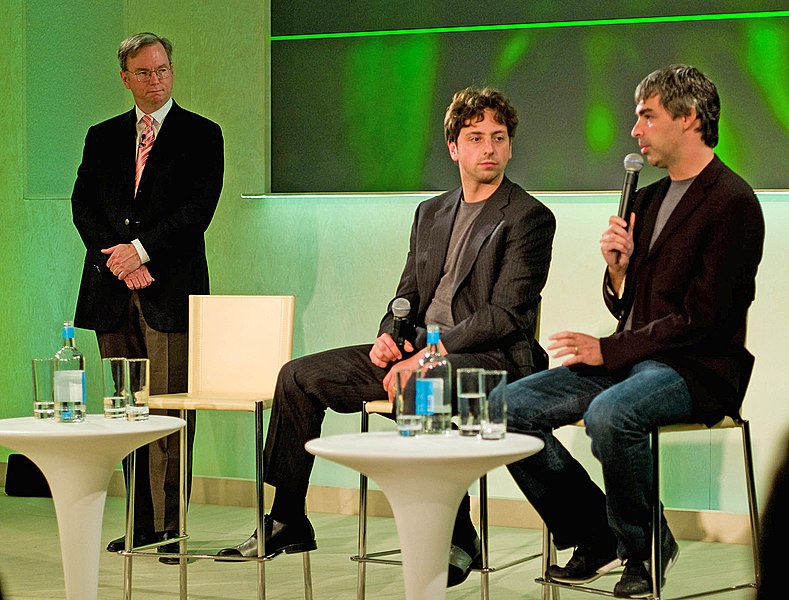 3 men on a stage wearing blazers, two sit on tall chairs speaking into microphones.