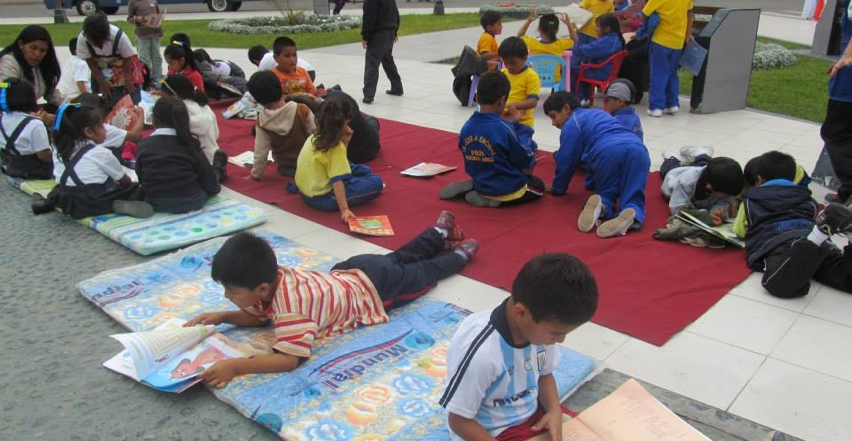 Young children sit and lie on colorful blankets on the ground, reading books and talking to adults.