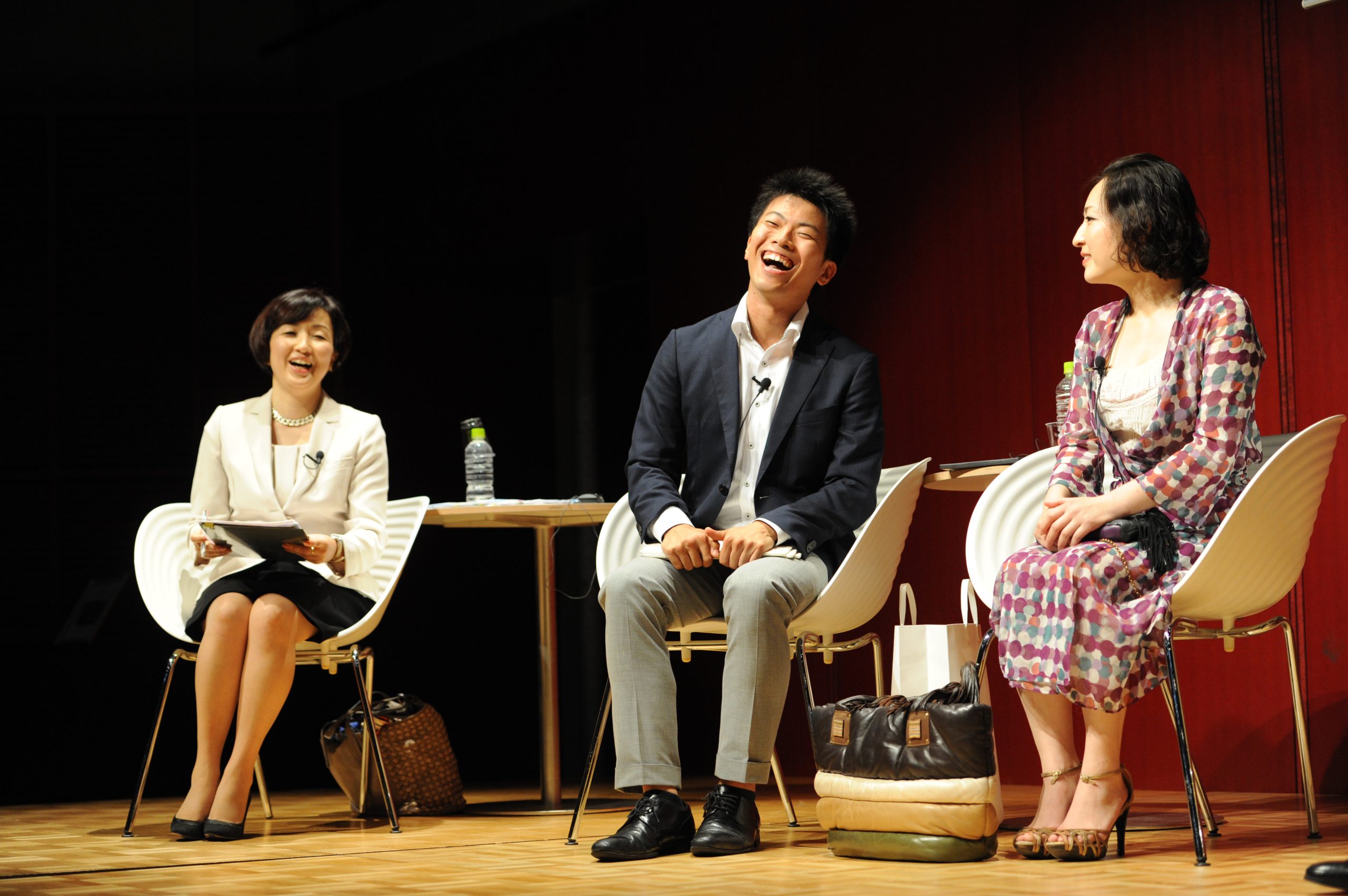 Two women in business dresses and a man in a blazer and slacks sit on a stage, laughing.