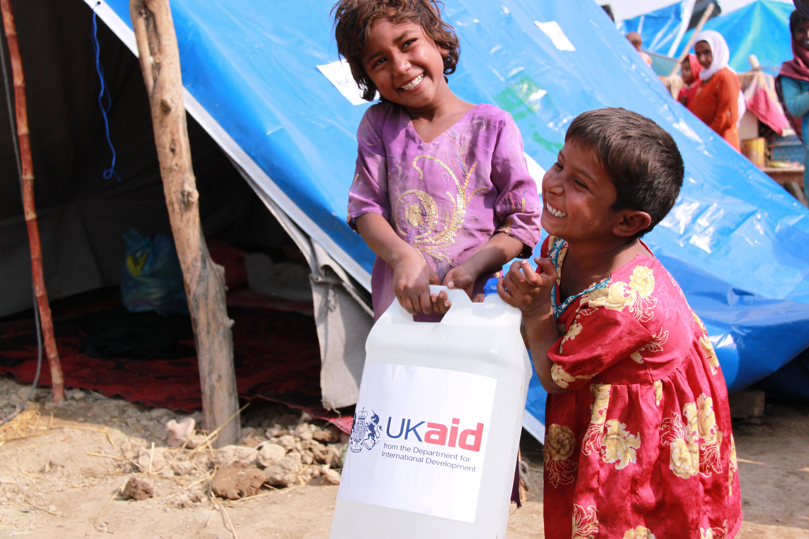 Two smiling young children in a refugee camp holding a UK aid bottle of water.