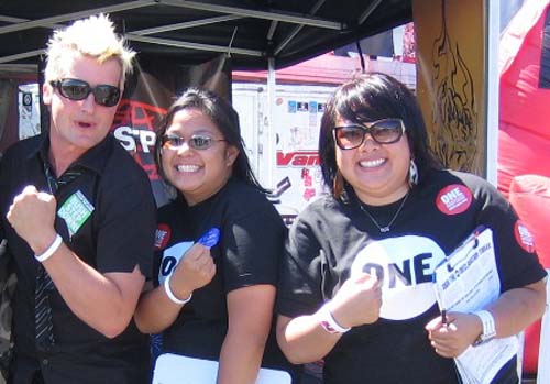 3 adults showing off their wrist bands, wearing "one" shirts and pins.